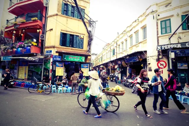 A busy road corner by day, a woman cycling with fruit baskets and plastic stools on the pavement
