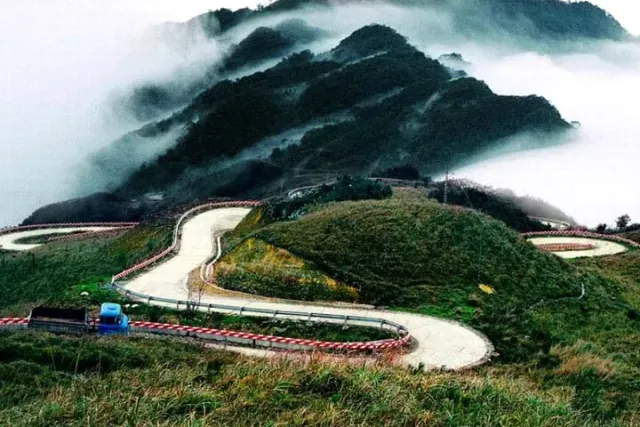 A winding road with red and white barriers around a hill, and cloud wrapped mountains