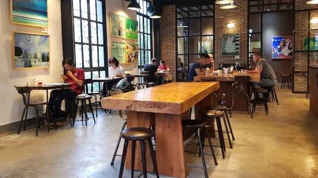 An industrial space with tall windows, a long shared wooden table, stools and people working