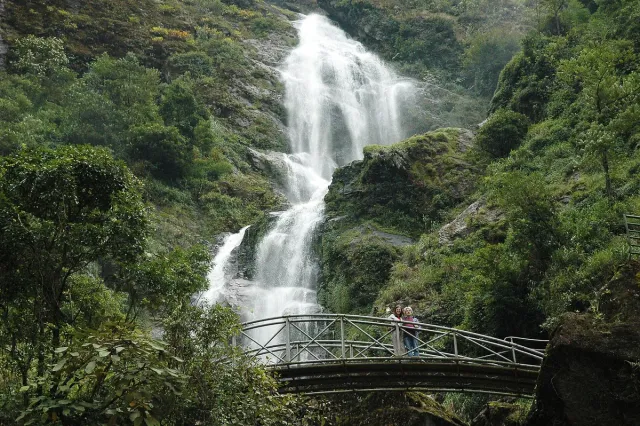 A tall cascade dropping in tiers down a green cliff, an arched metal bridge with visitors