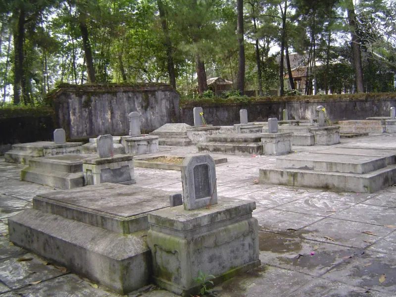 Rows of moss covered stone tombs in a paved walled yard inside the grounds of Tu Hieu pagoda, ringed by pine trees