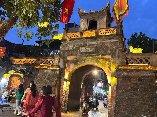 An old stone gate lit at night, motorbikes passing through the arch and passersby at the sides