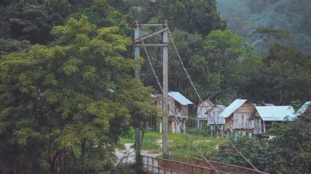 A suspension bridge pylon with cables, and behind it wooden stilt houses among trees
