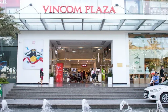 A white entrance with glass doors and steps, a jet fountain in front and shoppers coming out