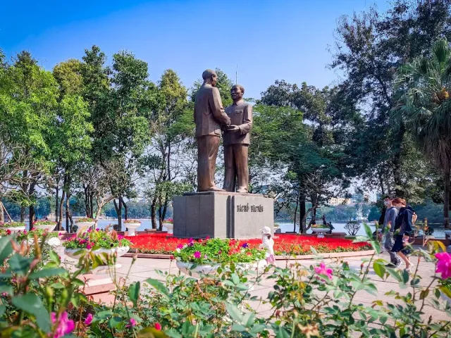 A bronze statue of two men shaking hands above flower beds, with a lake among the trees behind