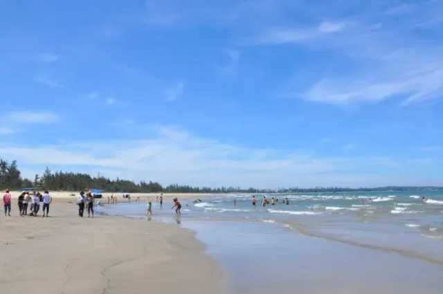 A wide flat strip of sand with groups of people at the waterline and casuarina trees behind