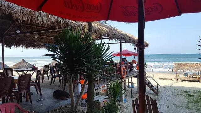 Tables under thatched roofs on the sand, facing a blue sea and swimmers on the beach