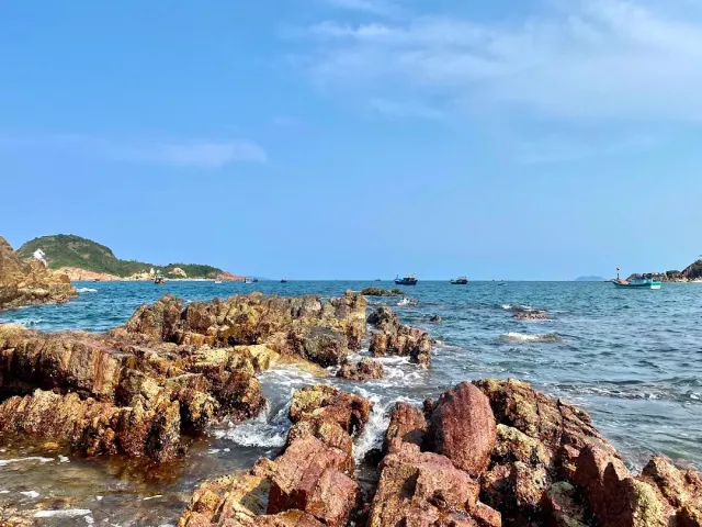 Jagged reddish rocks in the foreground, a blue sea with fishing boats and a headland
