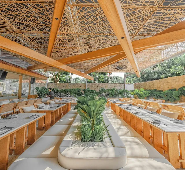 A hall under a woven wooden ceiling, with long tables and white padded benches