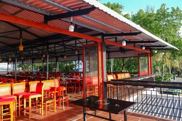 A terrace under a red metal roof with red and yellow stools and a table with a grill