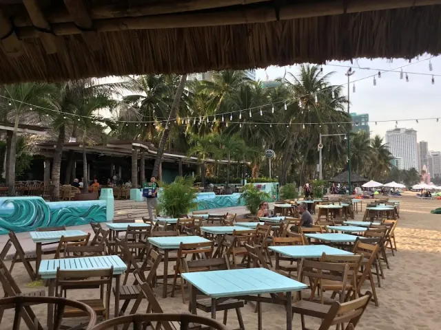 Wooden tables on the sand under a thatched roof, string lights and palms behind