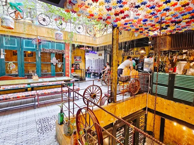 A crowded space with a colorful flower ceiling, bicycles and wooden wheels on the walls and an old cart