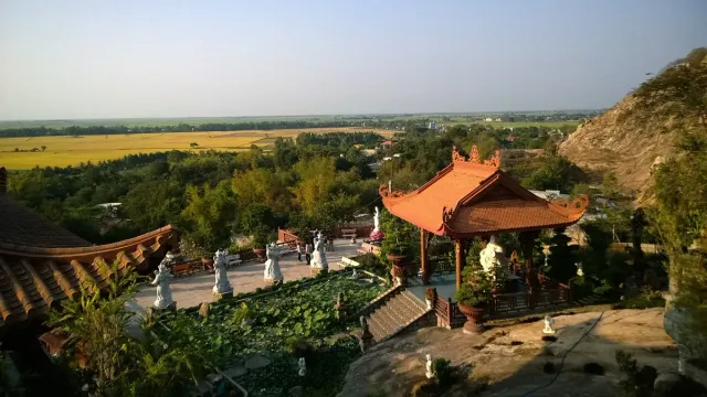 A hillside temple with an orange roofed pavilion, white statues along the path and cultivated plains below