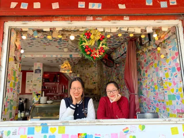 An open window whose walls are covered with thousands of colorful notes, two women leaning on the counter