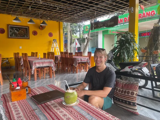 A young man sits before a coconut with a straw, behind him a dining hall with yellow walls and red tablecloths