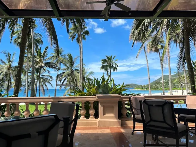 A covered terrace with dark armchairs and a stone balustrade, a view through palms to a turquoise sea