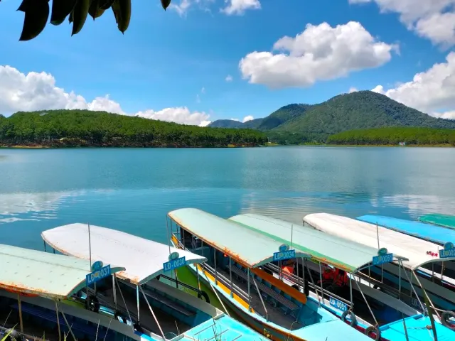 A row of moored tourist boats with colored canopies, calm water and forested hills