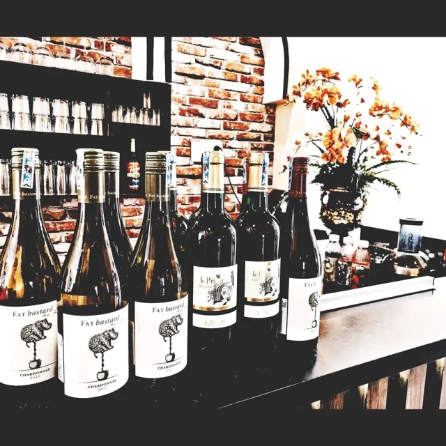 A row of white and red wine bottles on a dark counter, a brick wall and a flower vase behind