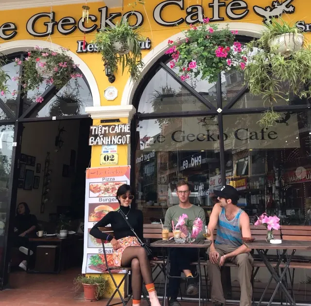 A yellow facade with large black letters, hanging flower baskets and three people seated outside