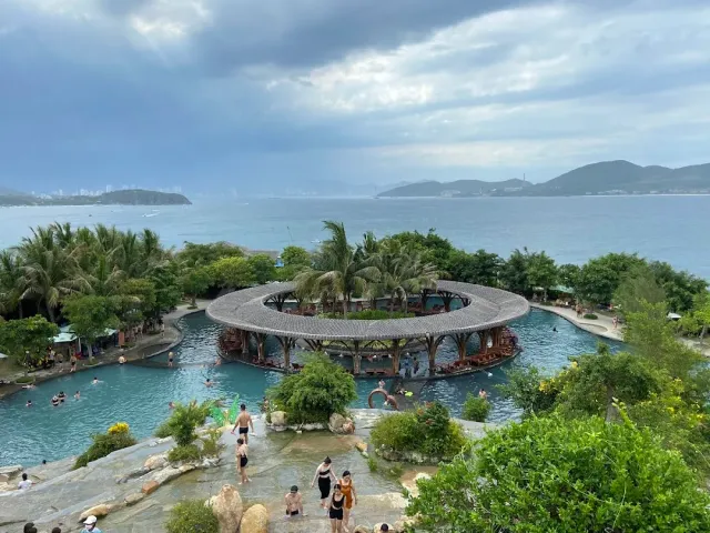 A pool complex with a round covered bar in the middle, bathers, palms and the sea behind