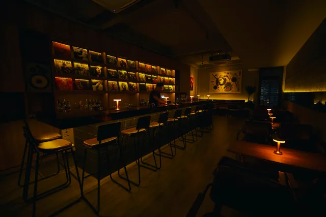 A dark bar with high stools and small table lamps facing a wall of record sleeves