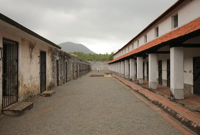 A long gravel yard between barred cell doors and a colonnade with red roof tiles