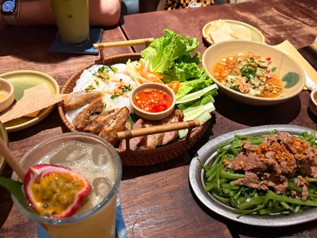 A wooden table with a straw tray holding lettuce and rolls, bowls of dishes and a glass of cold drink