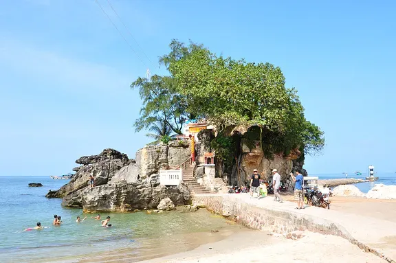 A low stone headland topped by a large tree and a small pavilion, with bathers in the water below