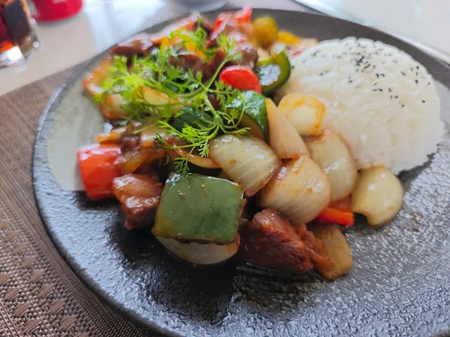 Stir fried meat with peppers and onion, beside white rice on a dark plate