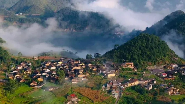 A view from above of tile roofed houses in a green basin, ribbons of mist on the mountains