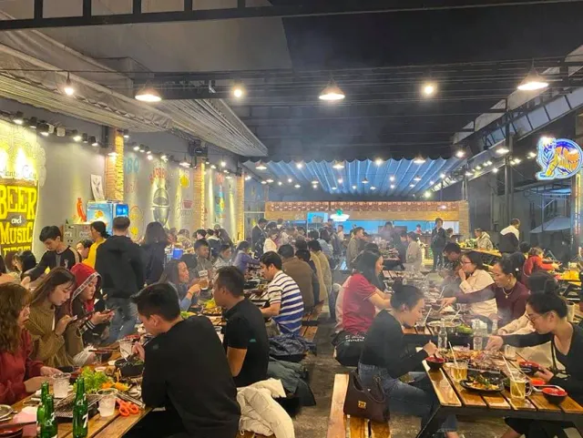 A bustling grill hall with smoke, neon signs and a crowd of diners along the tables