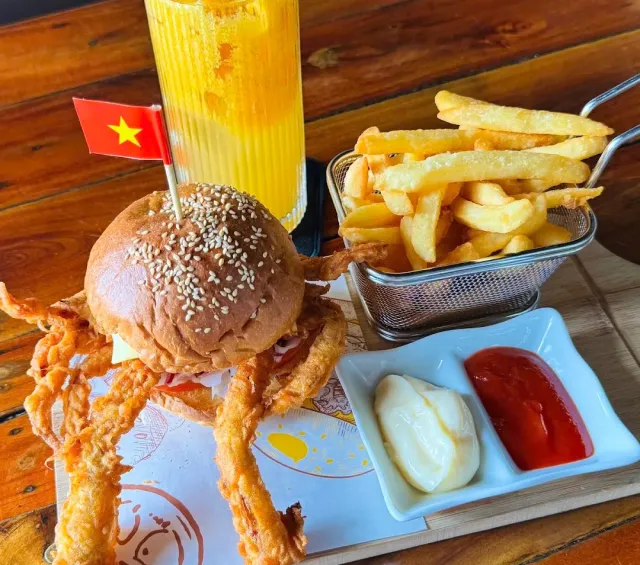 A soft shell crab burger with a small flag, a basket of fries and an orange drink on a wooden table