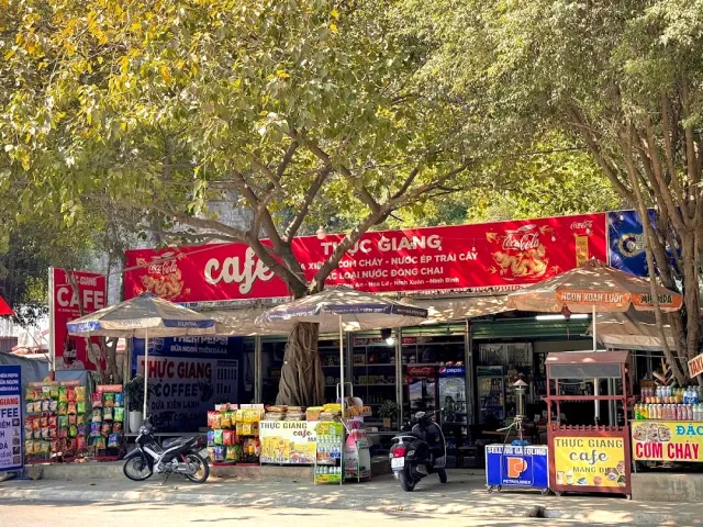 A roadside stand under big trees with a red sign, red parasols and parked scooters