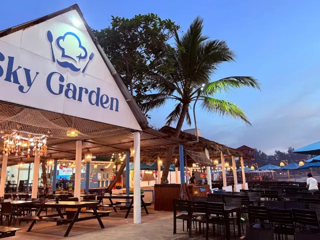 A building with a white gable and an illustrated sign, a netted roof, wooden tables and a tall palm