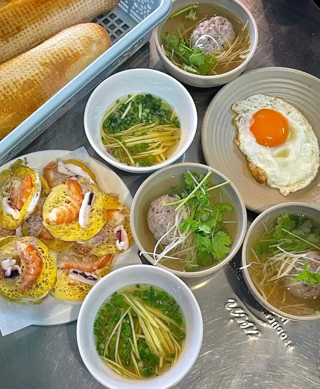 A metal counter with baguettes, pancakes topped with shrimp, bowls of broth and a fried egg