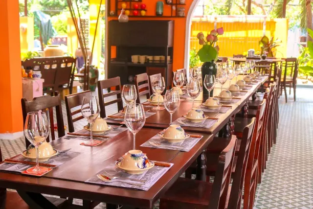 A long wooden table set with wine glasses and lidded bowls, orange columns in the background