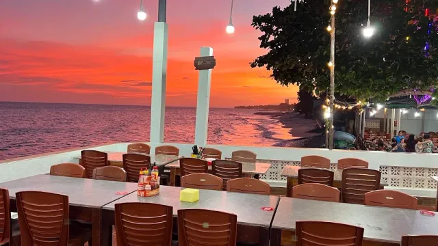 An orange sunset over the sea, metal tables and brown chairs on a terrace strung with bulbs