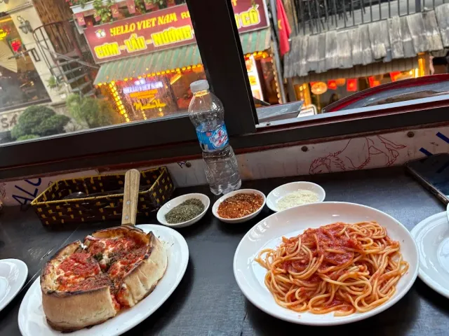 A deep dish pizza and pasta in tomato sauce on a table by a window facing the street