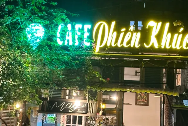 A night facade with a neon sign glowing green and yellow, tree branches in the foreground