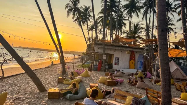 A sunset on the beach with colorful beanbags on the sand, palm trunks and a string of lights