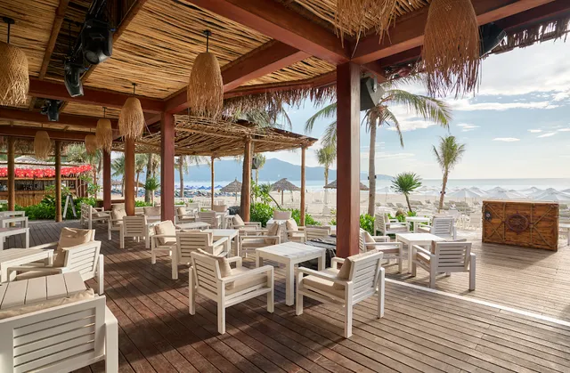 A thatch covered wooden deck with white chairs, facing a beach with parasols and mountains