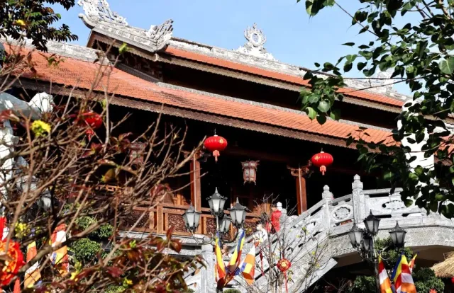 A wooden hall with an orange tiled roof and red lanterns, a stone bridge and small flags in front