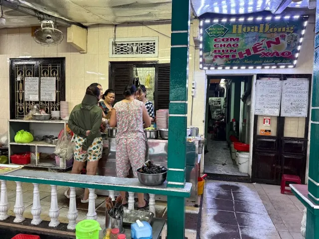 An alley stall with a lit sign, women preparing food at the counter and bowls stacked ready