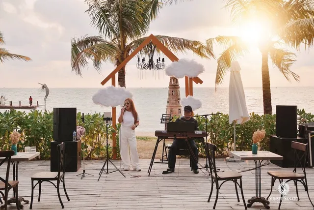 A singer and a keyboard player perform on a wooden deck facing the sea and palms at sunset