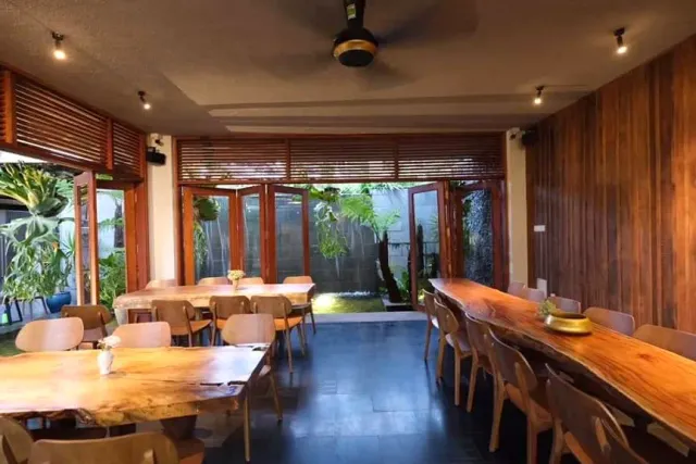 A wide space with two long wooden tables, a dark floor and louvered doors onto a green courtyard