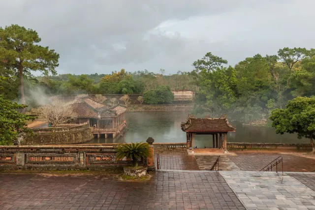 A grey tomb complex on the shore of a lake surrounded by forest
