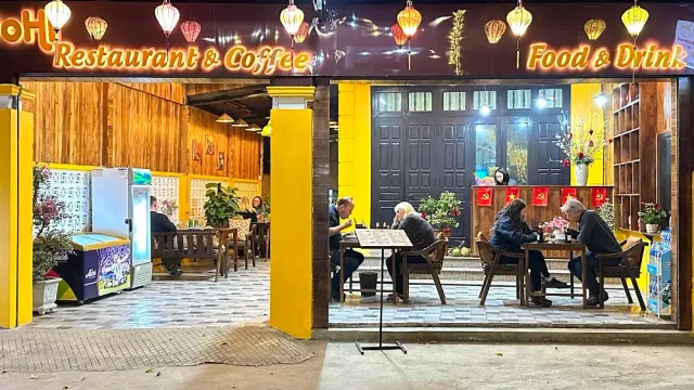 A night facade with yellow pillars, a lit sign, lanterns and guests seated outside