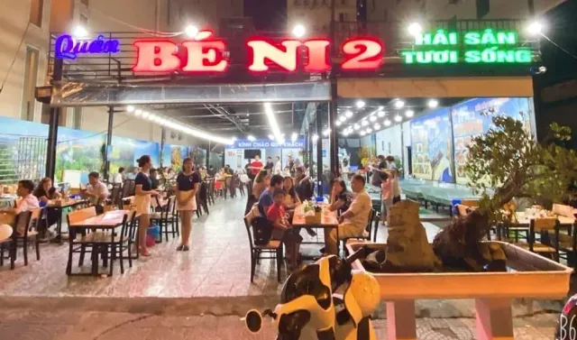 A large red neon sign above an open seating area with diners in the evening