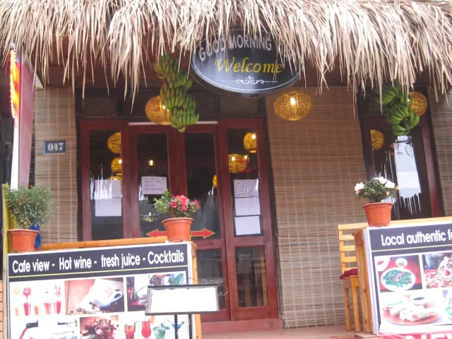 A front with a thatched roof, an oval welcome sign, hanging banana bunches and woven lanterns