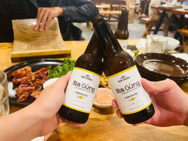 Two hands clinking beer bottles over a wooden table with a plate of grilled meat